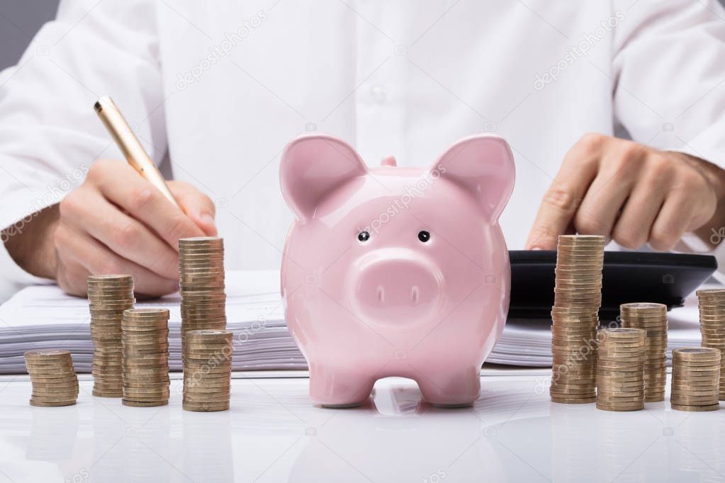 Close-up Of Businessman Calculating Bill With Piggybank And Stacked Coins On Desk In Office