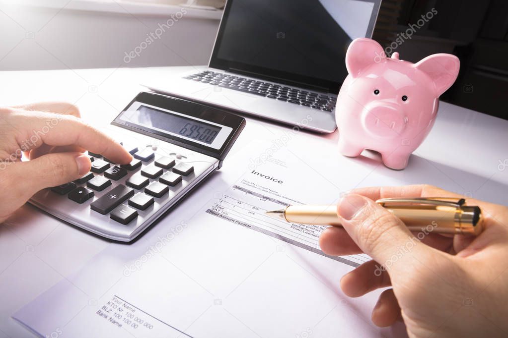 Businessperson's Hand Calculating Invoice With Calculator And Piggybank And On Desk In Office