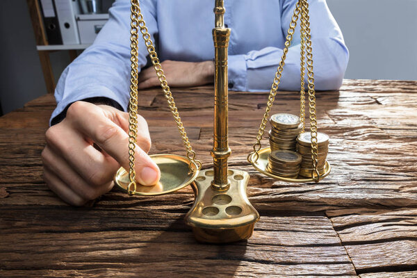 Close-up Of A Businessperson Balancing Stacked Coins On Justice Scale With His Finger