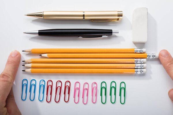 Person 's Finger Arranging The Pencils with Row Of Pins Rubber and Pen On White Background
