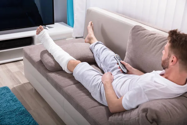 Mladý Muž Zlomenou Nohou Ležel Pohovce Drží Vzdálené Poblíž Televize — Stock fotografie