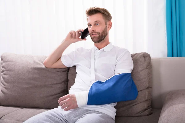 Young Man With Fractured Hand Sitting On Sofa Talking On Mobile Phone