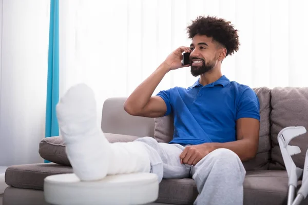 Jovem Feliz Com Perna Quebrada Sentado Sofá Conversando Telefone Móvel — Fotografia de Stock