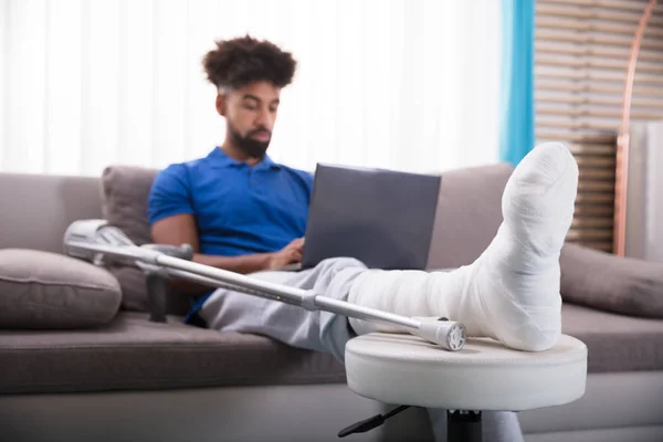 用手提电脑坐在沙发上的男子断腿 — 图库照片