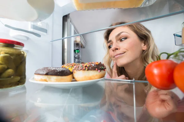 关闭一个困惑的年轻女子在打开冰箱看甜甜圈 — 图库照片