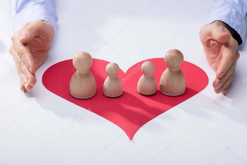 Human Hand Protecting Wooden Pawns On Red Heart Over White Background