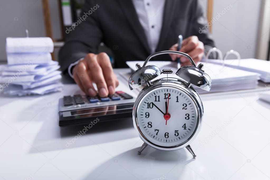 Businessperson Using Calculator For Calculating Bill With Alarm Clock On Desk