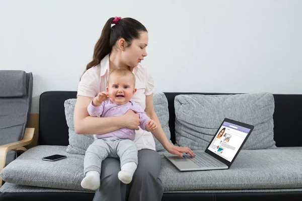 Woman With Her Crying Baby Chatting On Social Networking Site Using Laptop