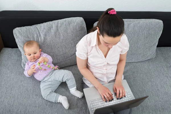 Kobieta Siedzi Kanapie Przy Użyciu Laptopa Podczas Jej Dziecko Bawi — Zdjęcie stockowe