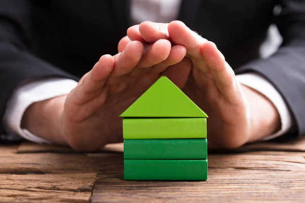 关闭一个生意人的手保护木桌上的绿色房子模型 — 图库照片