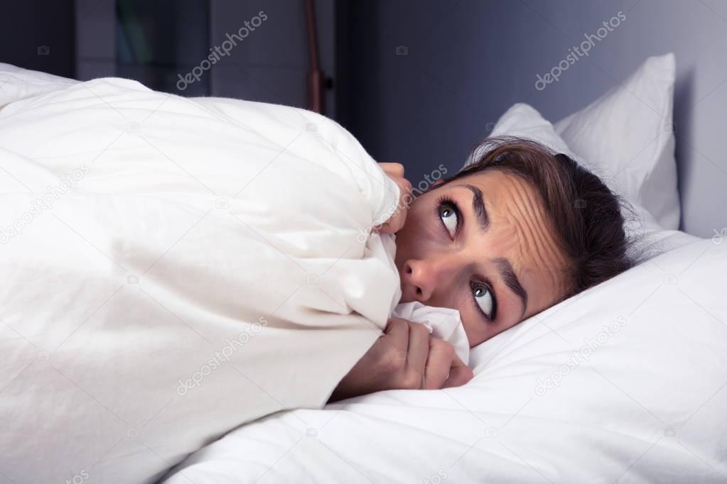 Scared Woman Pulling Bed Sheet Over Self On Bed At Night