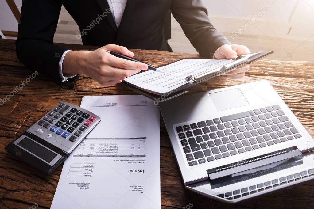Businessperson Analyzing Document On Clipboard With Invoice And Laptop On Wooden Desk
