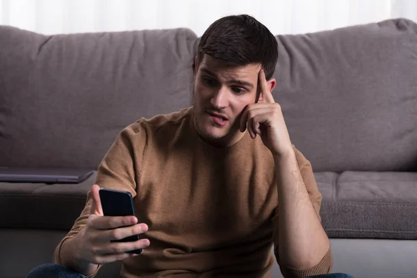 Martwi Się Młody Człowiek Patrząc Smartphone Siedząc Kanapie Domu — Zdjęcie stockowe