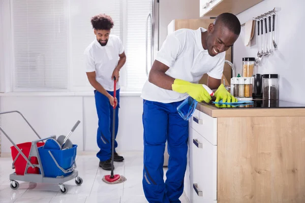 Two Smiling Male Janitor Cleaning The Induction Stove And Mopping Floor In The Kitchen