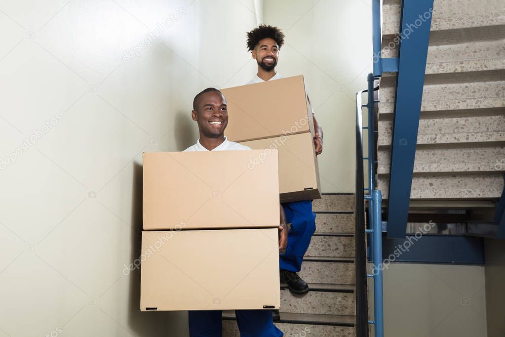 Portrait Of Two Young Smiling Movers Standing On Staircase Holding Cardboard Boxes