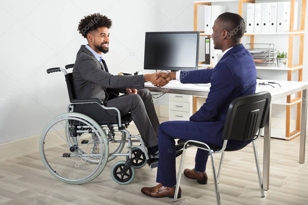 Two Happy Businessmen Sitting On Wheelchair And Chair Shaking Hands In Front Of Computer