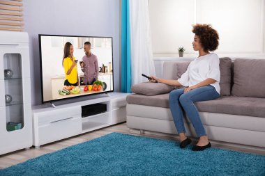 Evde televizyon izlerken uzaktan tutarak koltukta oturan genç kadın