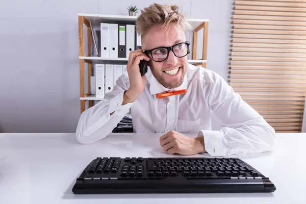 机の上のキーボードで携帯電話で話している幸せな青年実業家 — ストック写真
