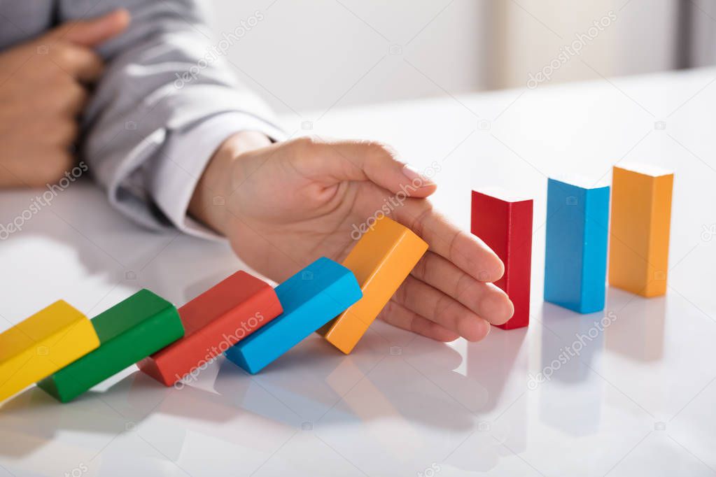 Close-up Of Businessperson Hand Stopping Colorful Blocks From Falling On Table In Office