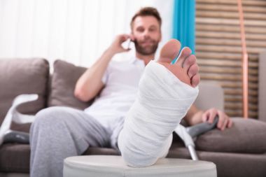 Kırık bacak Smartphone'da kanepe konuşmak oturan ile mutlu genç adam
