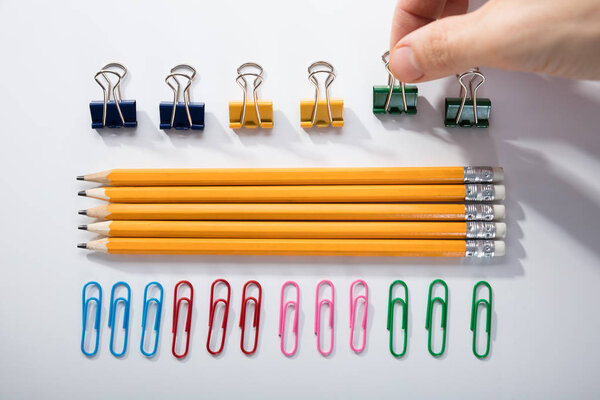 Person 's Finger Arranging The Pencils with Row Of Pins Rubber and Pen On White Background
