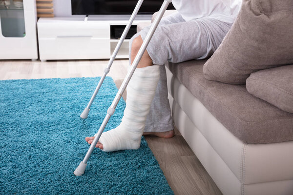 Young Man With Broken Leg Using Crutches To Get Up From Sofa