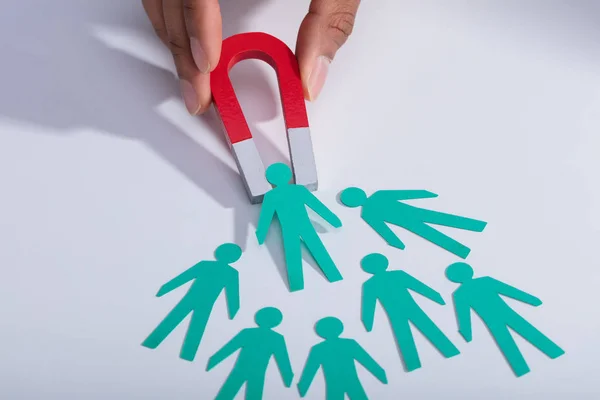 Close-up Of A Businessperson's Hand Holding Red Horseshoe Magnet Attracting Paper Candidates On Desk
