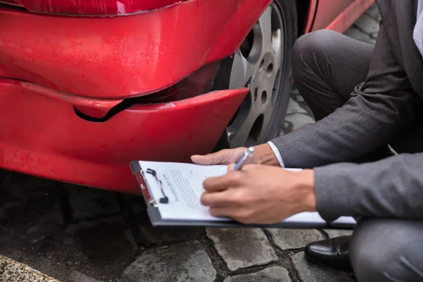 破損した赤い車を調べながらクリップボードに若い男性の保険エージェント執筆 — ストック写真