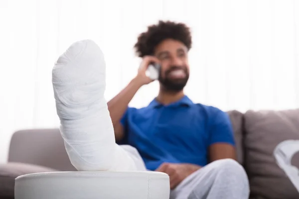 Szczęśliwy Młody Mężczyzna Złamaną Nogą Siedząc Kanapie Rozmowy Telefon Komórkowy — Zdjęcie stockowe