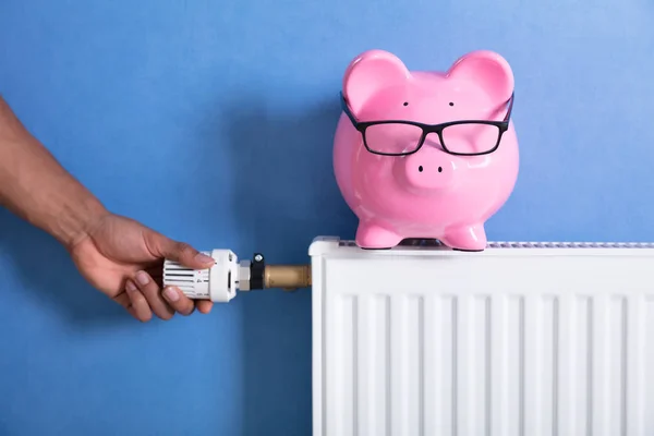 一个人的手调节恒温器与小猪银行散热器 — 图库照片