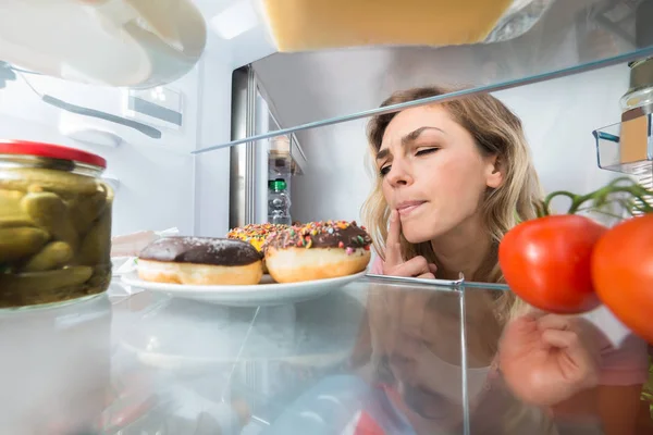 关闭一个困惑的年轻女子在打开冰箱看甜甜圈 — 图库照片