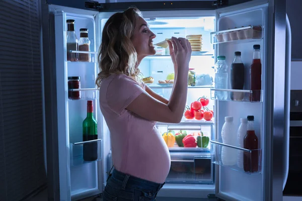 一个年轻的孕妇在冰箱前吃一片奶酪 — 图库照片