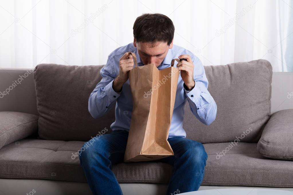 A Man Sitting On Sofa Vomiting Into Paper Bag At Home