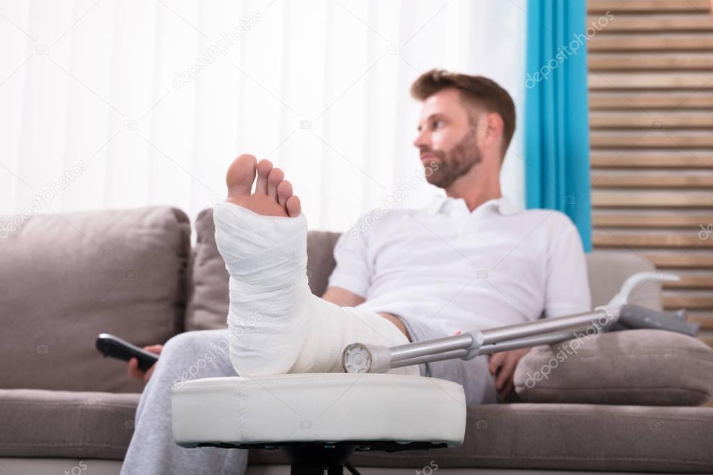 Young Man With Broken Leg Sitting On Sofa Holding Remote