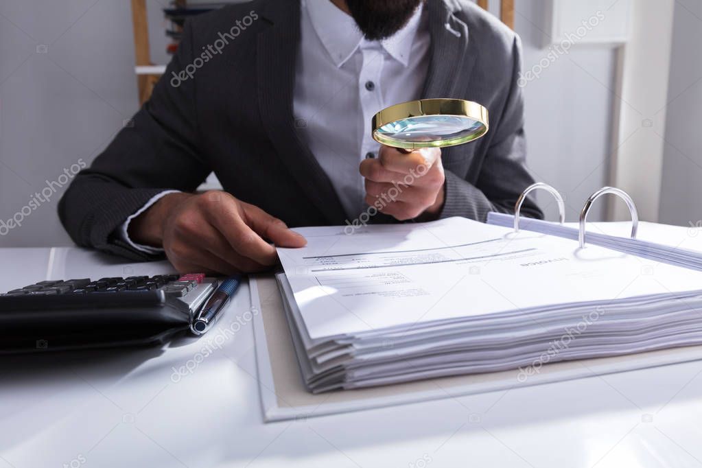 Businessperson's Hand Analyzing Bill With Magnifying Glass At Workplace