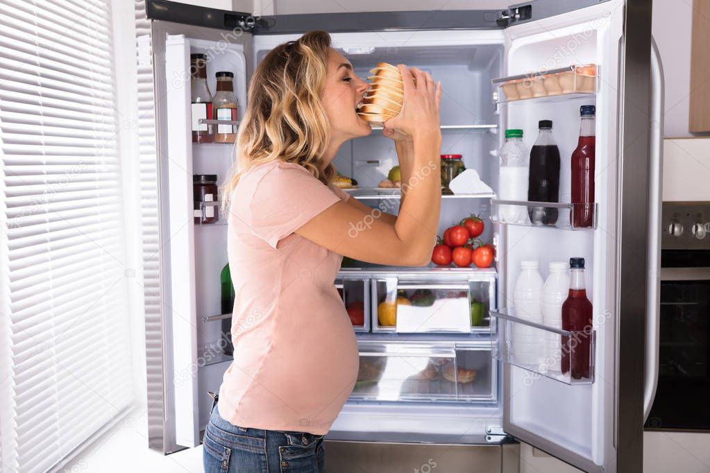 Pregnant Woman Eating Sandwich In Front Of Open Refrigerator