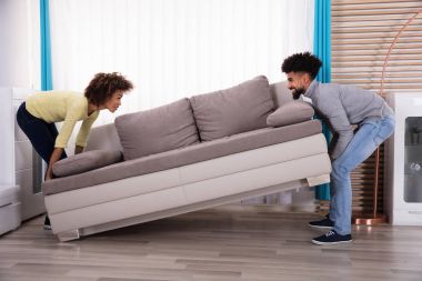 Side View Of Young Smiling Couple Lifting Sofa In Living Room