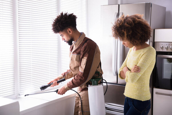 Женщина смотрит на молодого работника по борьбе с вредителями, распыляющего инсектицид на подоконнике
