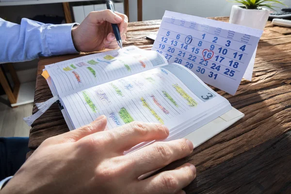 Businessperson's Hand Checking Schedule In Diary With Calendar On Wooden Desk