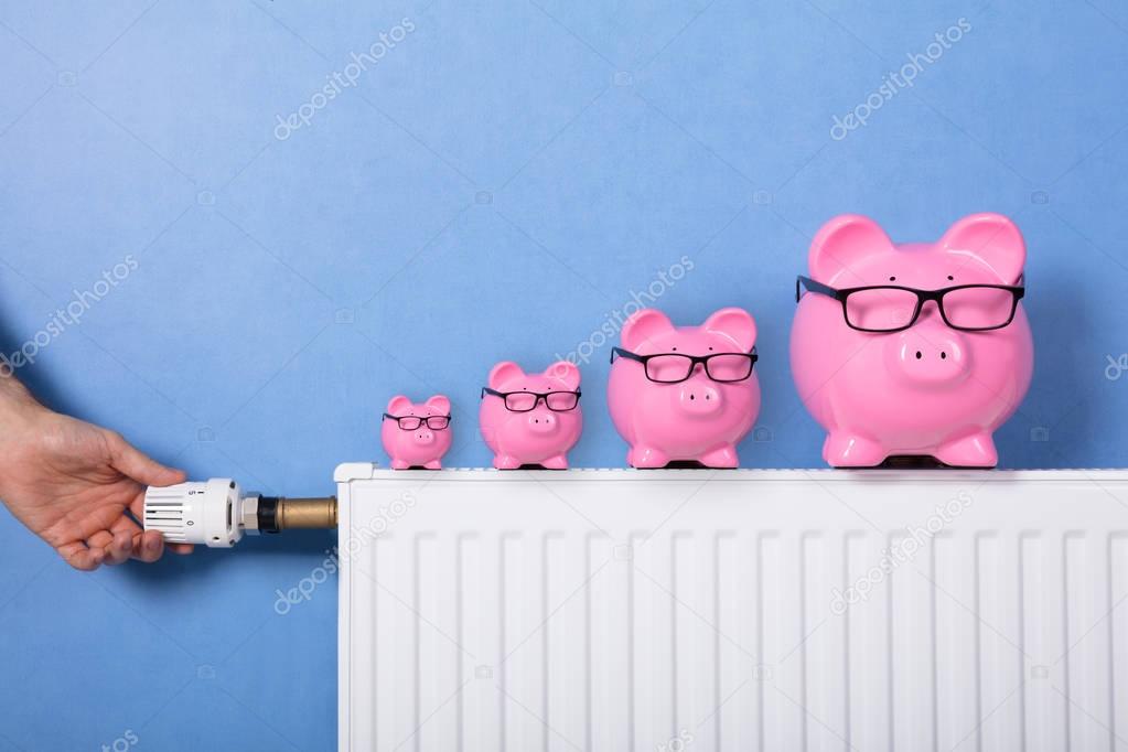Close-up Of A Person's Hand Adjusting Thermostat With Piggy Banks Wearing Eyeglasses On Radiator