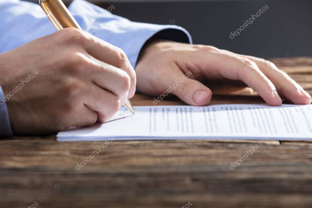 Close-up Of A Businessperson's Hand Signing Document On Wooden Desk