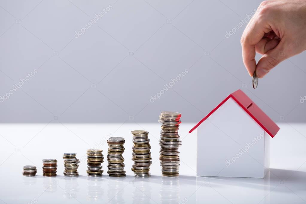 Person's Hand Inserting Coin In The House Model With Increasing Coins Stack On Desk