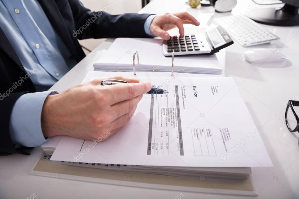 Close-up Of A Businessperson's Hand Checking Invoice With Calculator At Workplace