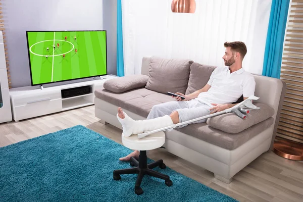坐在沙发上看足球比赛在电视上的年轻人断腿 — 图库照片