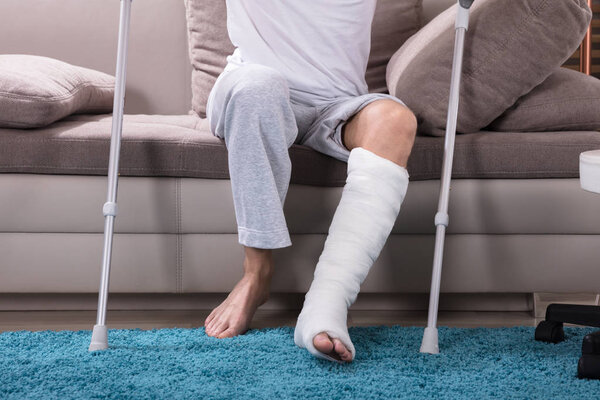 Young Man With Broken Leg Using Crutches To Get Up From Sofa