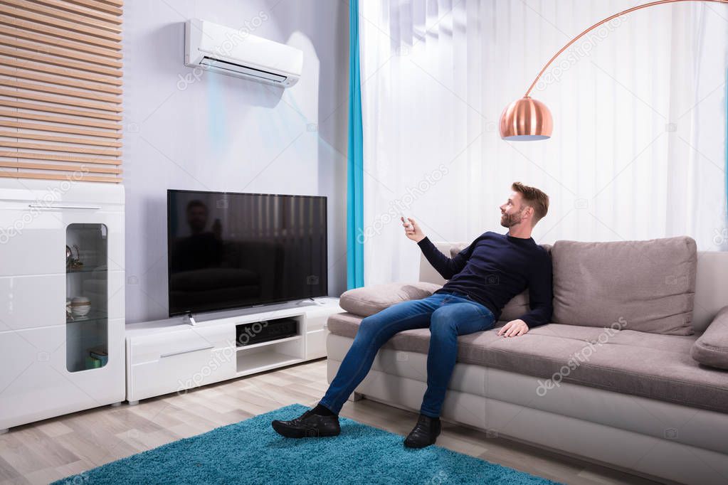 Young Man Sitting On Sofa Using Air Conditioner At Home