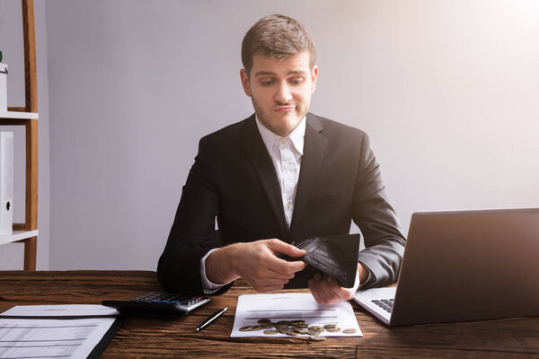 Sad Businessman Checking His Wallet With Coins And Laptop On Wooden Desk