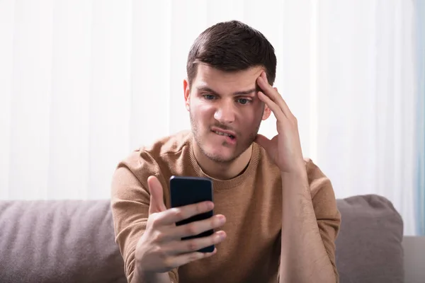 Hombre Joven Preocupado Mirando Teléfono Inteligente Sentado Frente Sofá Casa —  Fotos de Stock