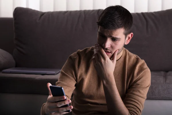 Jovem Preocupado Olhando Para Smartphone Sentado Frente Sofá Casa — Fotografia de Stock