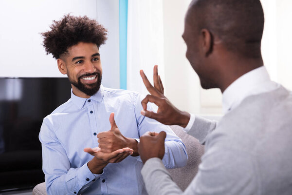 Two Young Happy Men Sitting On The Sofa Making Sign Languages